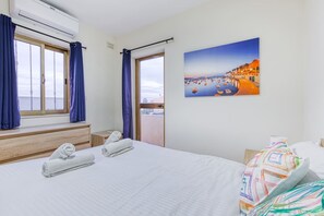 A hotel room with a bed, a window with curtains, an air conditioning unit, a painting of a coastal scene, and a view of the sea. - St James the Traditional 4-bed Townhouse Terrace (Sliema)