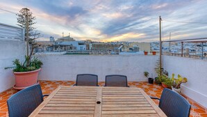 Una terraza en la azotea con una mesa y sillas de madera, plantas en macetas y vistas de la ciudad.