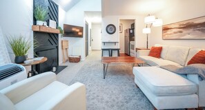 Una sala de estar moderna con un sofá blanco, una mesa de centro y una chimenea. Hay un televisor montado en la pared y un cuadro sobre el sofá.