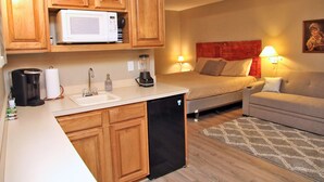 A compact kitchen with wooden cabinets, a microwave, and a dishwasher. There is a bed with a red headboard and a beige sofa in the adjacent room. - Shanty Creek 2bd 2ba Adjoining Rooms Ski-golf (Mancelona)