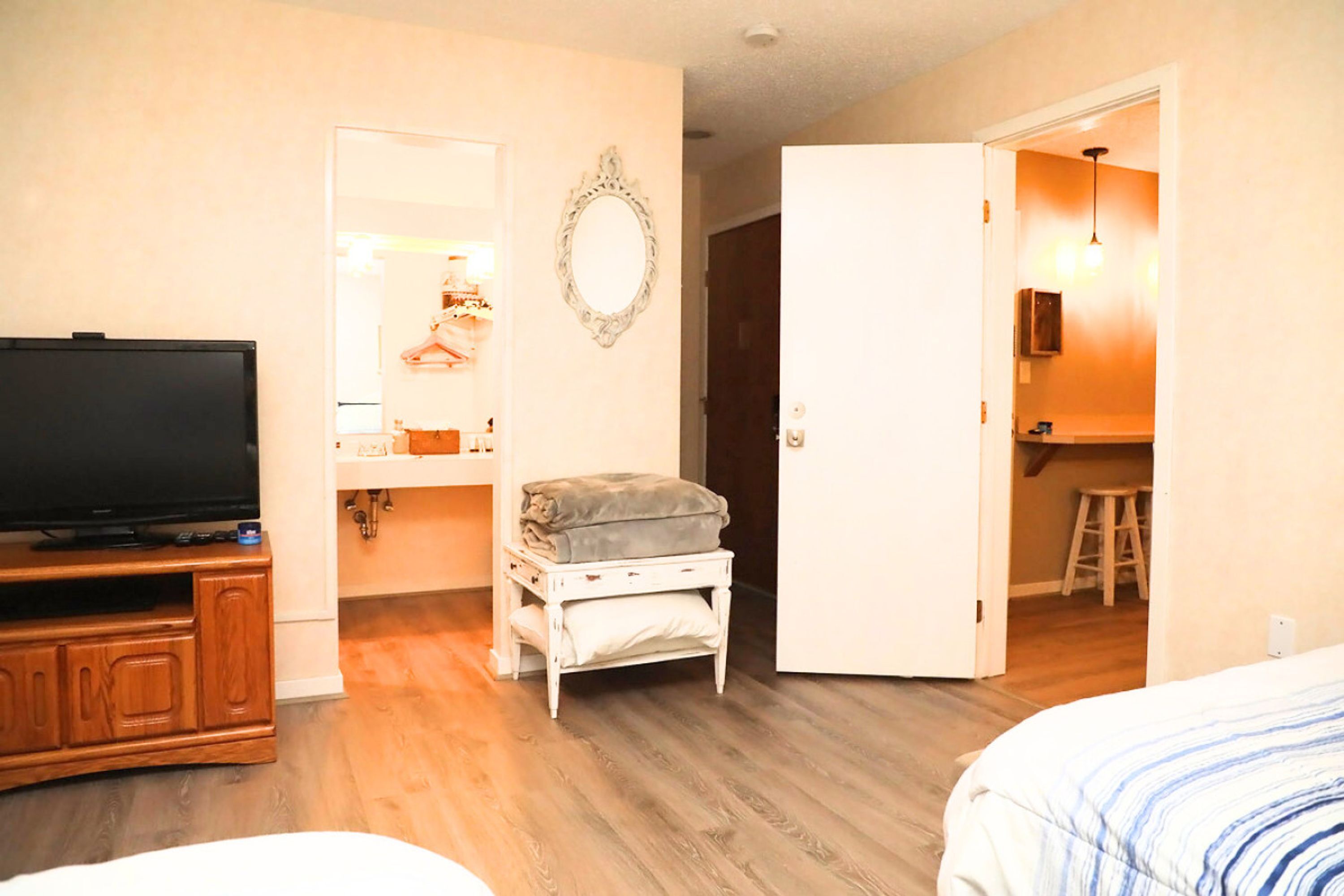 A hotel room with a television on a wooden cabinet, a mirror on the wall, a small table with a folded blanket, and a bed with a striped blanket.