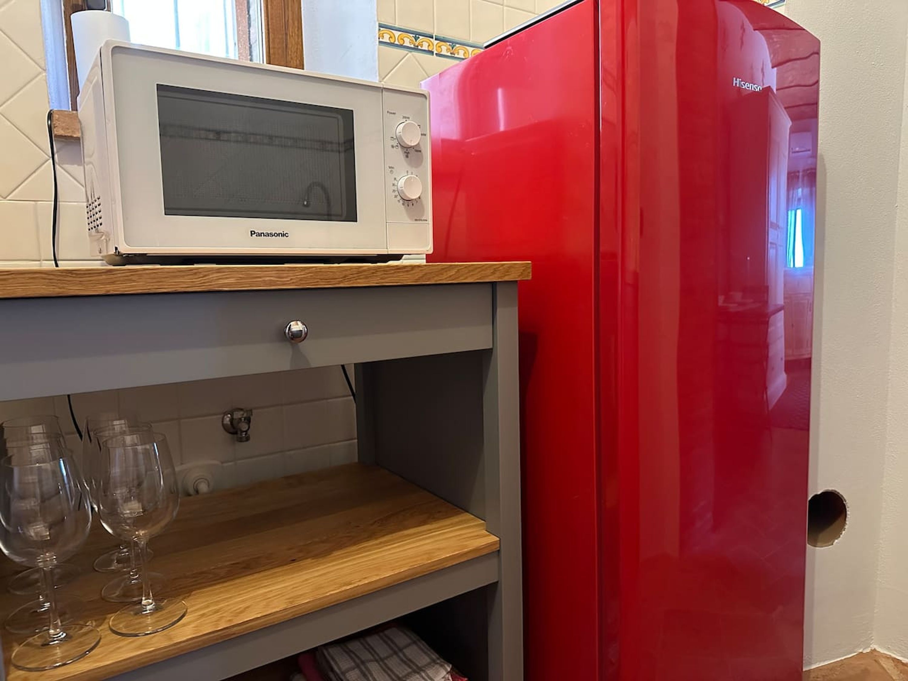 Una cocina con un microondas sobre un estante de madera, un refrigerador rojo y una copa de vino en un estante inferior.