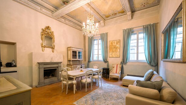 Un salon spacieux doté d’une cheminée, d’un lustre, d’une table à manger avec des chaises, d’un canapé et d’une grande fenêtre avec des rideaux.