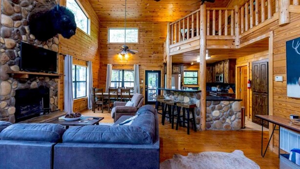 A log cabin interior with a stone fireplace, a large sofa, a dining area, and a kitchen with a stone countertop.