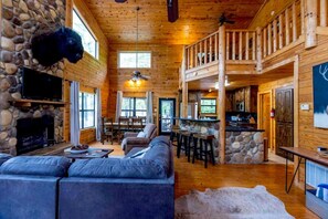 A log cabin interior with a stone fireplace, a large sofa, a dining area, and a kitchen with a stone countertop. - Bison Point Smoker Grill Corn Hole Fire Pit (Broken Bow)