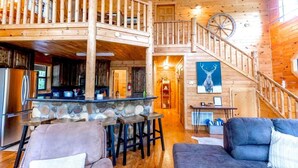 A spacious cabin interior with a stone fireplace, a dining area, a living room with a sofa, and a staircase leading to an upper deck.