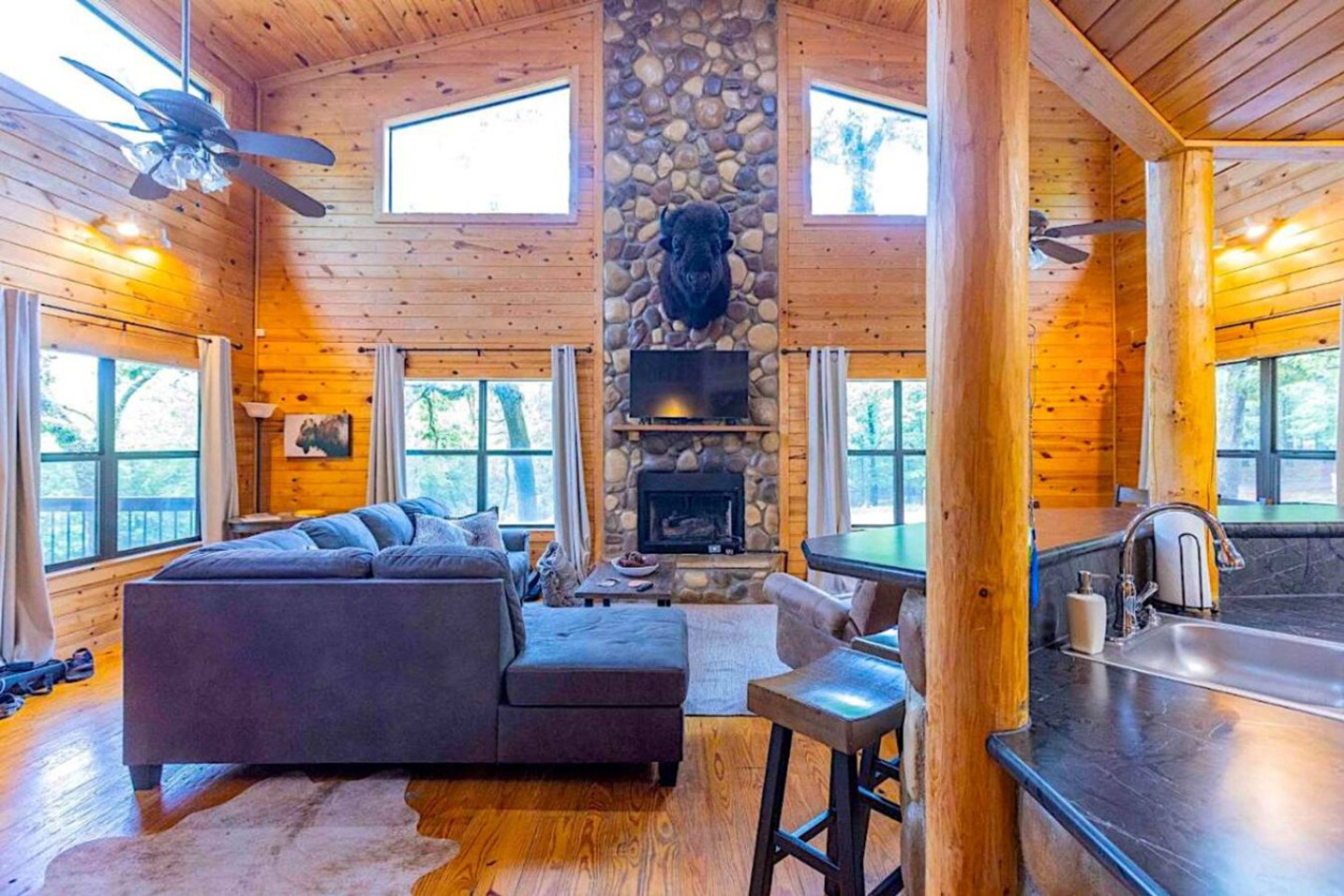 A cozy cabin interior with a stone fireplace, wooden walls, a sofa, and a bar area.