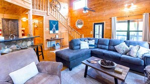 A spacious living room with a large sectional sofa, a fireplace, a dining area with a stone bar, and a staircase leading to an upper level.