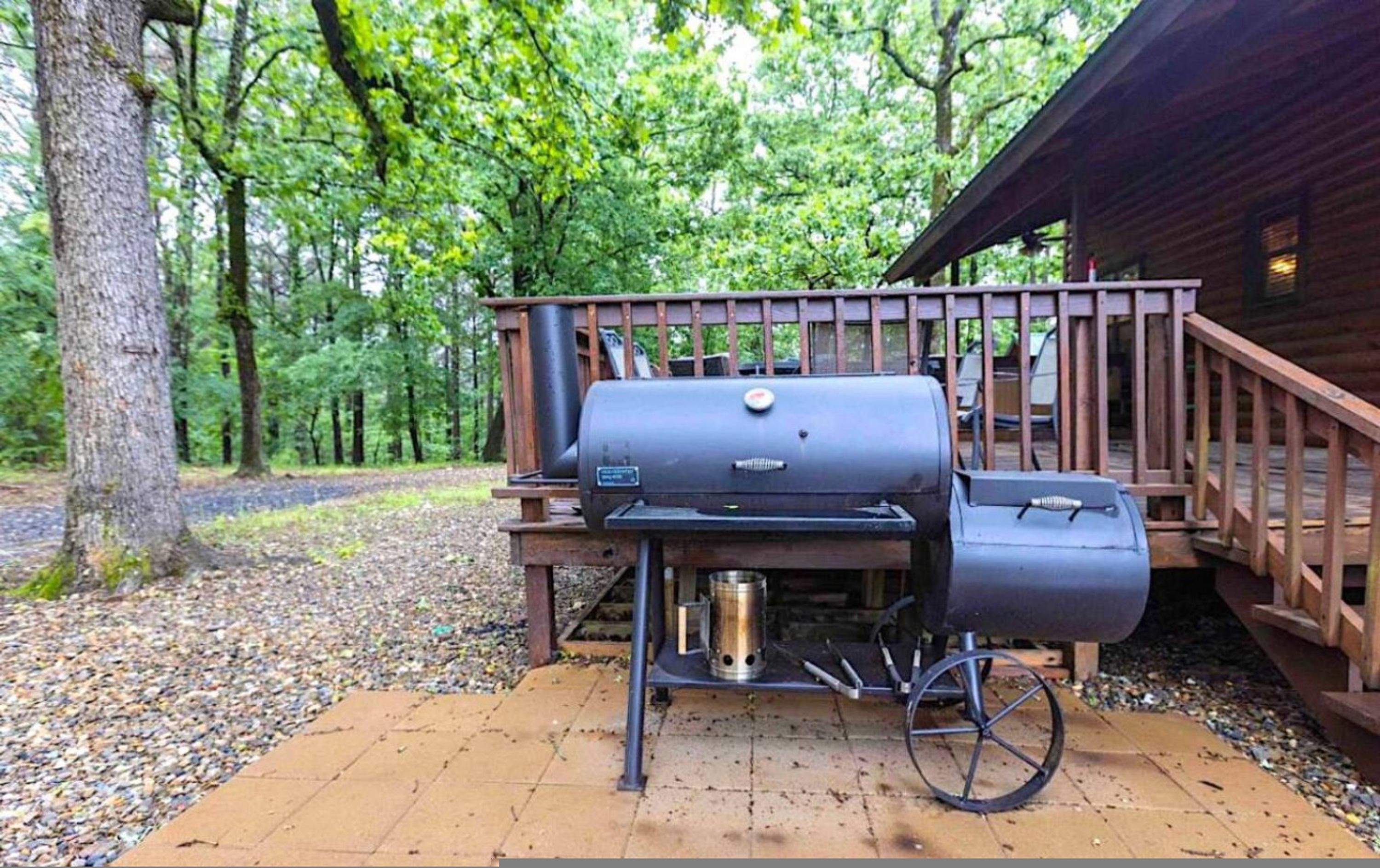A portable grill set up on a wooden deck with a metal wheelbarrow and a metal container nearby.