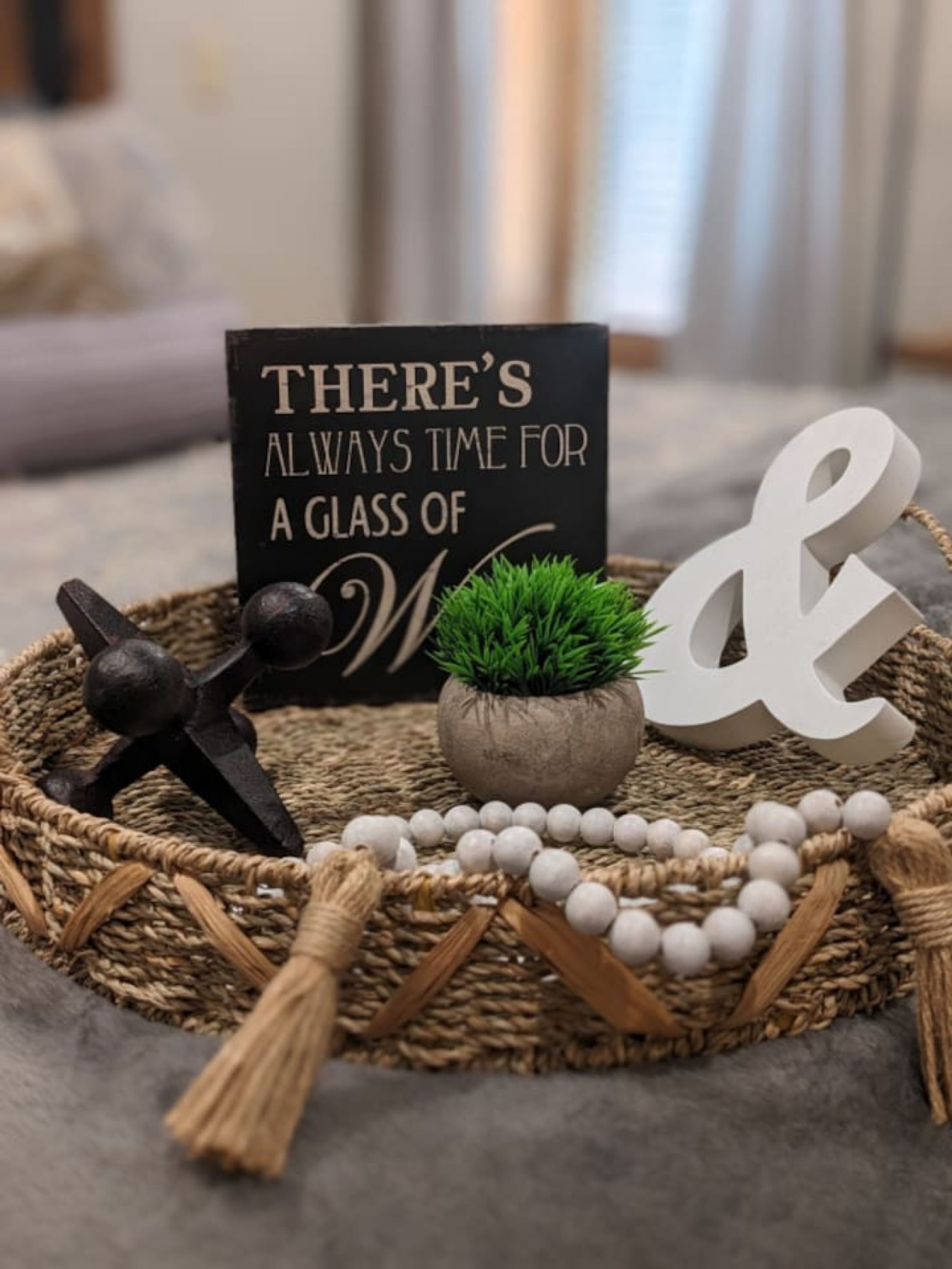Eine dekorative Anordnung mit einem Schild, das "THERE'S ALWAYS TIME FOR A GLASS OF M" zeigt, einem kleinen Topfpflanzchen, einem dekorativen Buchstaben 'M' und einer weißen Perlenkette.