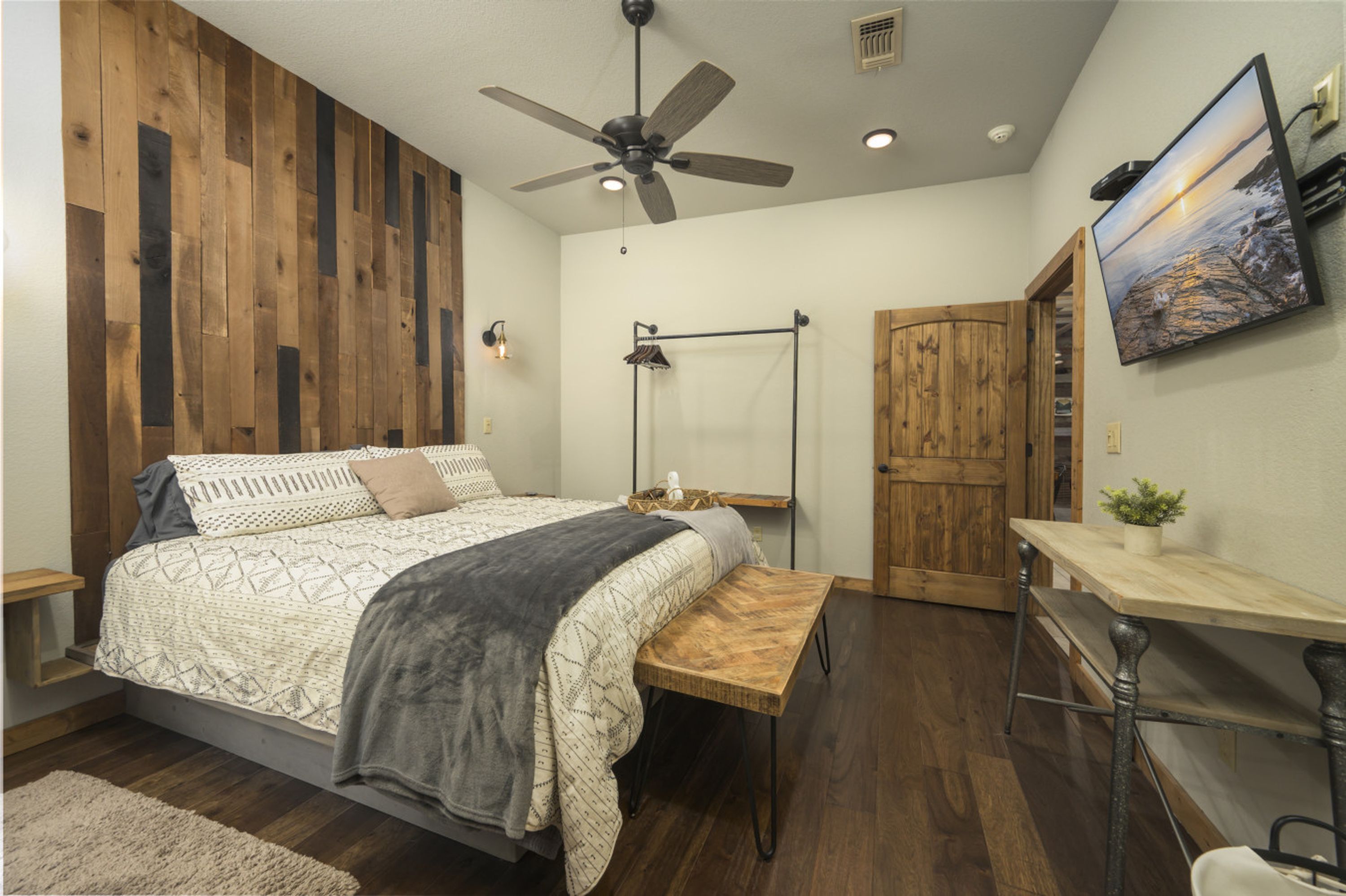Een slaapkamer met een houten wand, een bed met beddengoed met een patroon, een plafondventilator, een nachtkastje, een houten bankje, een bureau, een stoel, een aan de muur bevestigde televisie en een deur.