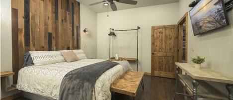 Een slaapkamer met een houten wand, een bed met beddengoed met een patroon, een plafondventilator, een nachtkastje, een houten bankje, een bureau, een stoel, een aan de muur bevestigde televisie en een deur.