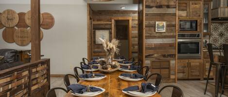 Una mesa de comedor de madera dispuesta con platos y servilletas, rodeada de sillas, en una habitación con paredes de madera y un candelabro rústico.