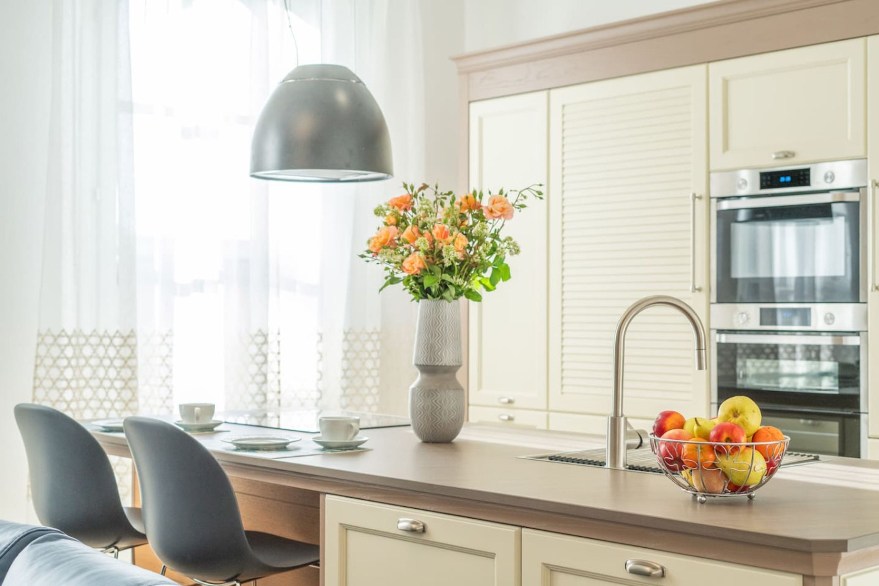 A modern kitchen with a dining area, featuring a sink, a vase with flowers, and a fruit bowl.
