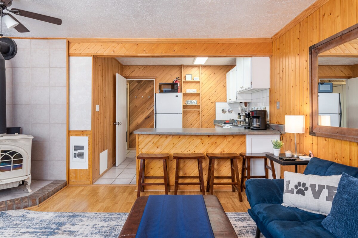 A compact living space with a kitchenette, a dining area with wooden stools, a blue sofa with a 'LOVE' pillow, and a small rug.