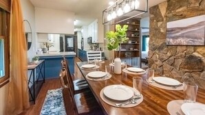 A dining area with a long wooden table set for four, a stone accent wall, and a view into the kitchen. - Blue Ridge Gateway Cabin With Mountain Views (Moravian Falls)