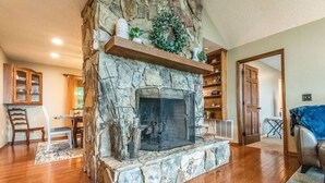 A stone fireplace with a mantel shelf, wooden flooring, and a dining area with a table and chairs. - Blue Ridge Gateway Cabin With Mountain Views (Moravian Falls)
