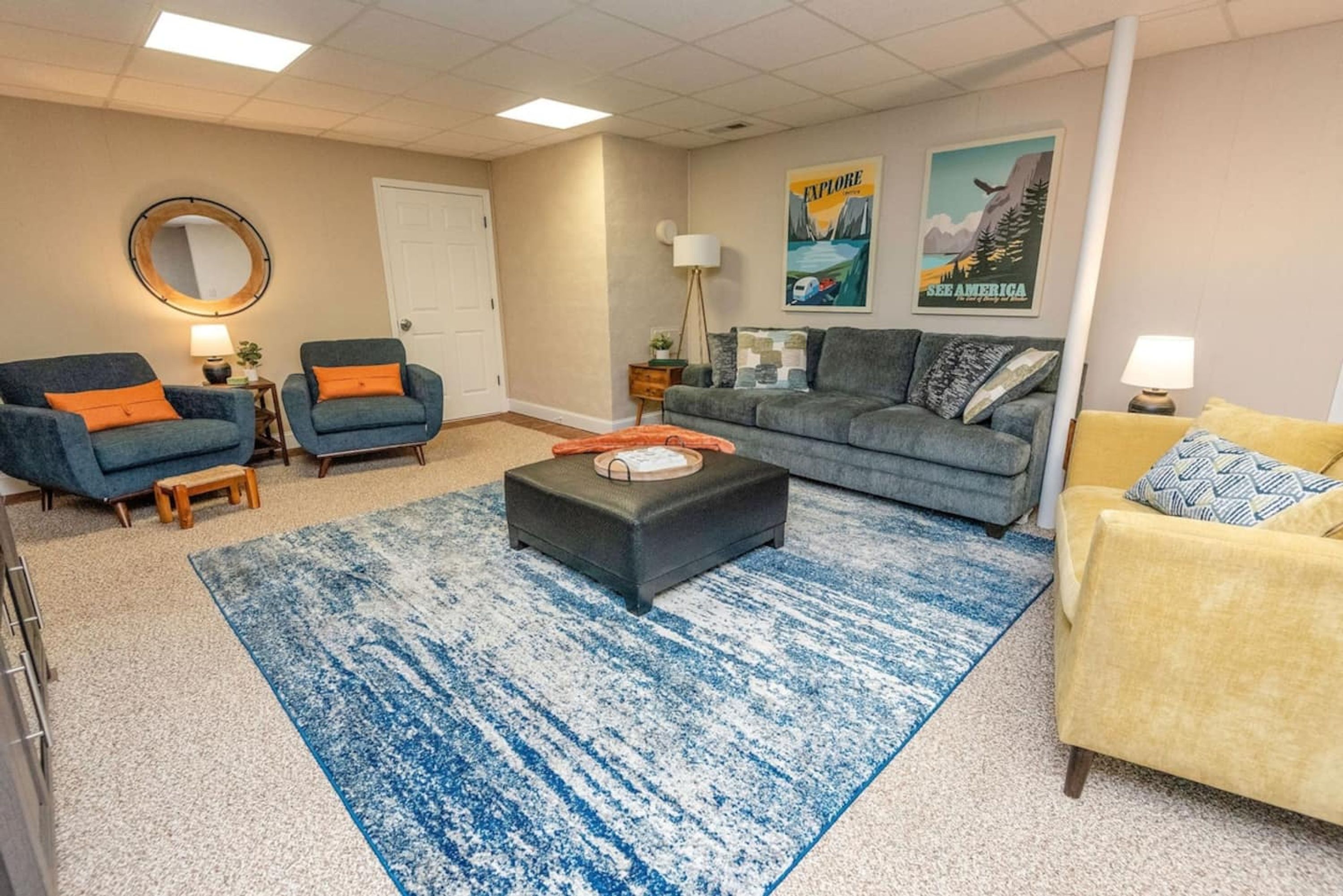 Een woonkamer met een grijze bank, twee blauwe fauteuils, een gele fauteuil, een ronde spiegel, een lamp, een poster en een blauw vloerkleed.
