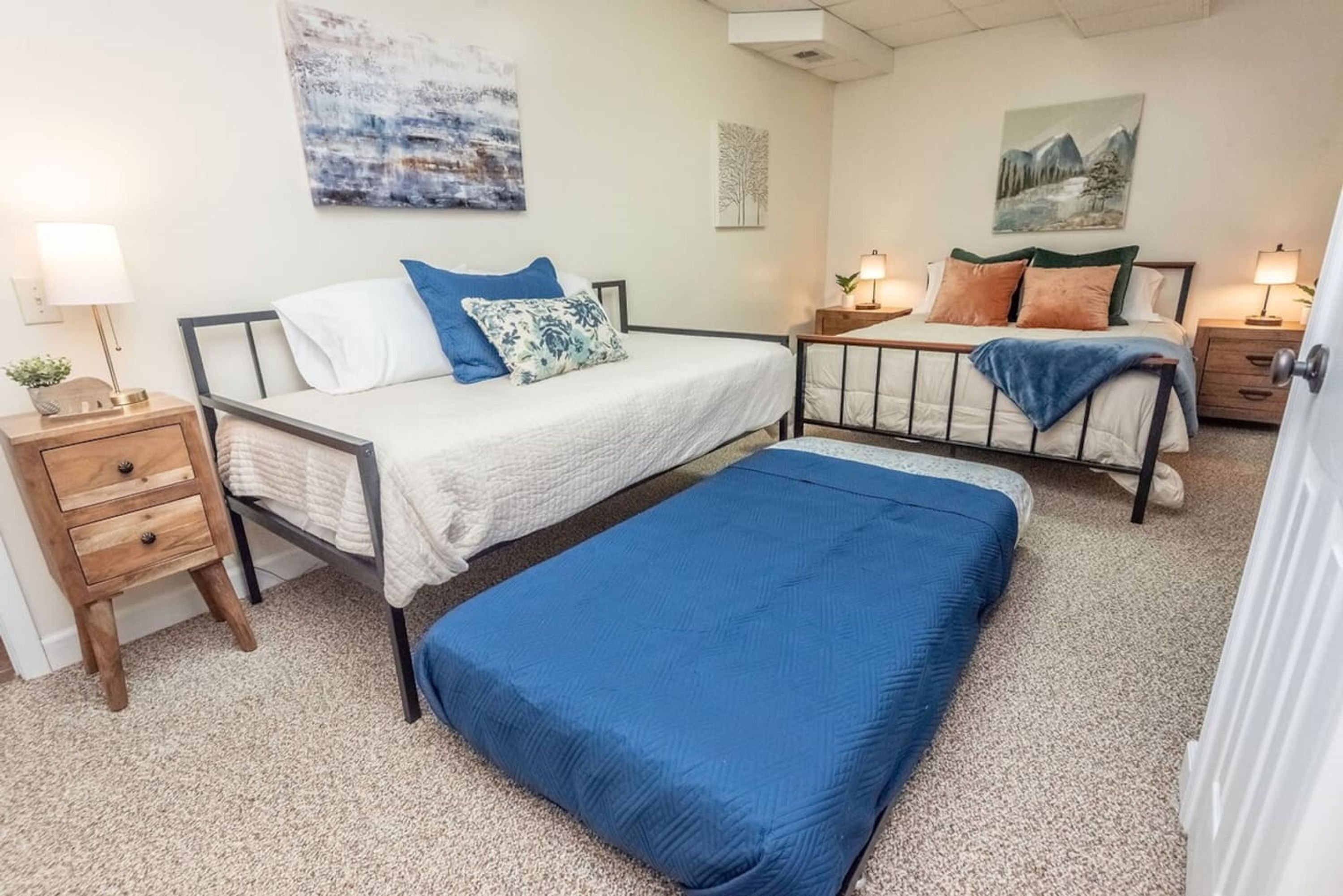 A bedroom with two beds, each with a nightstand, a lamp, and a painting on the wall.