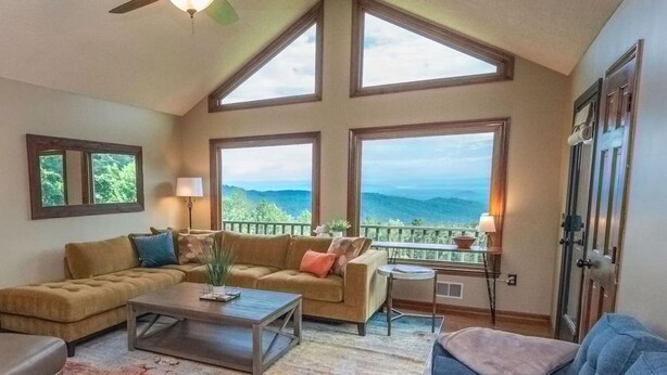A spacious living room with a high ceiling, large windows offering a mountain view, a ceiling fan, and a cozy seating area with a sofa and armchairs. - Blue Ridge Gateway Cabin With Mountain Views (Moravian Falls)
