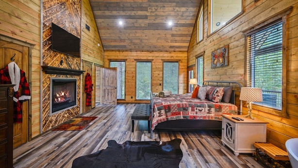 Um quarto de cabana rústica com lareira, uma cama com roupa de cama estampada, um banco e uma janela com persianas.