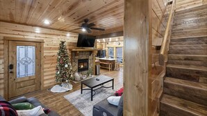 A cozy log cabin interior with a stone fireplace, a Christmas tree, a television, and a staircase. - Three Bears Hideout Luxury Mountain Cabin Getaway for Families Groups (Almont)
