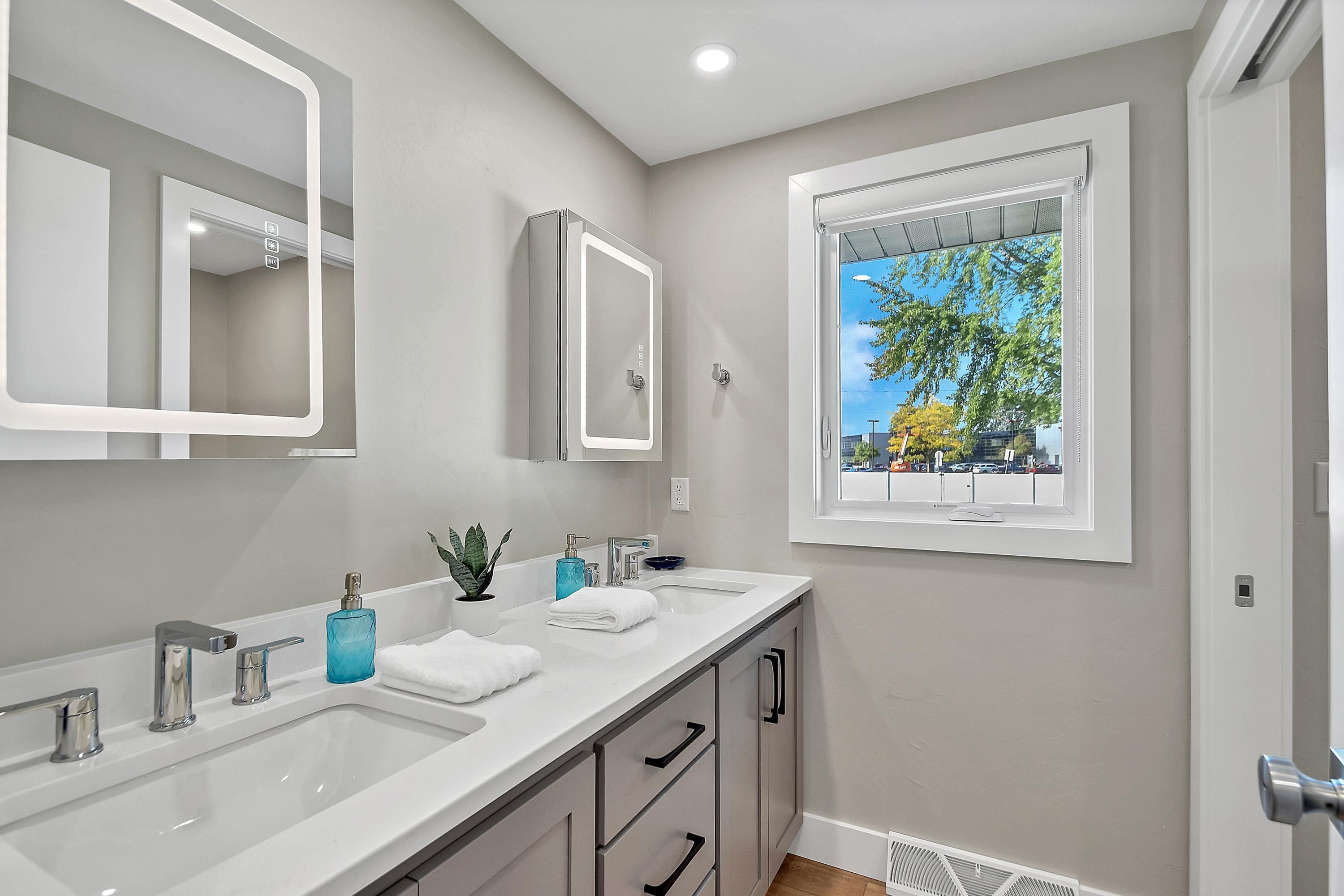 A modern bathroom with two sinks, a large mirror, and a window with a view of trees and buildings.