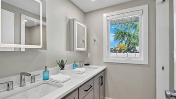 A modern bathroom with two sinks, a large mirror, and a window with a view of trees and buildings.