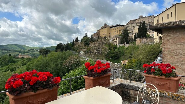 A terrace with a table and chairs, potted red flowers, and a view of historic buildings in the background.