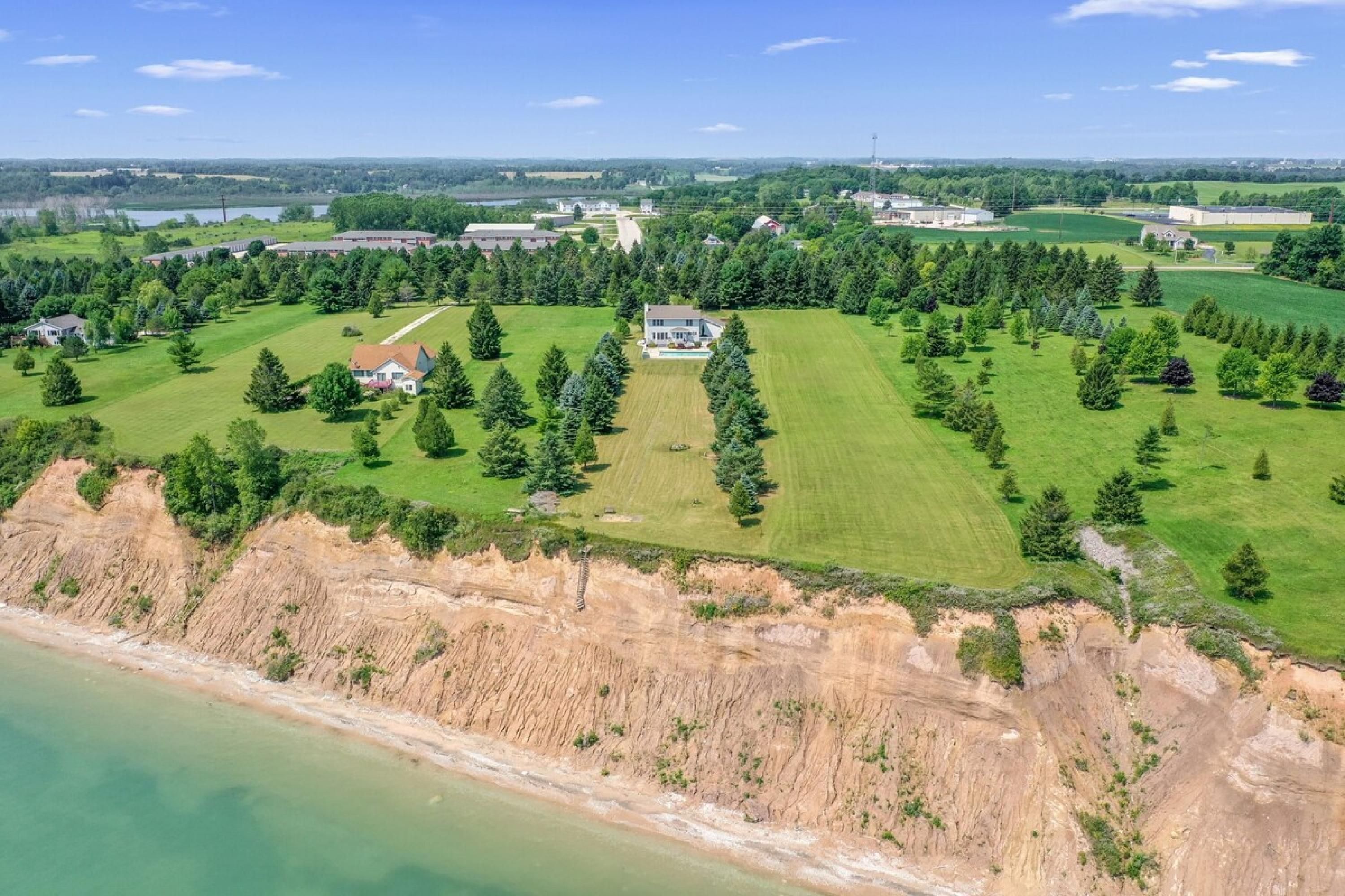 Luchtbeeld van een landgoed aan de kust, met een groot huis omgeven door groene velden en bomen, een strand en een verre uitzichting over een stad.