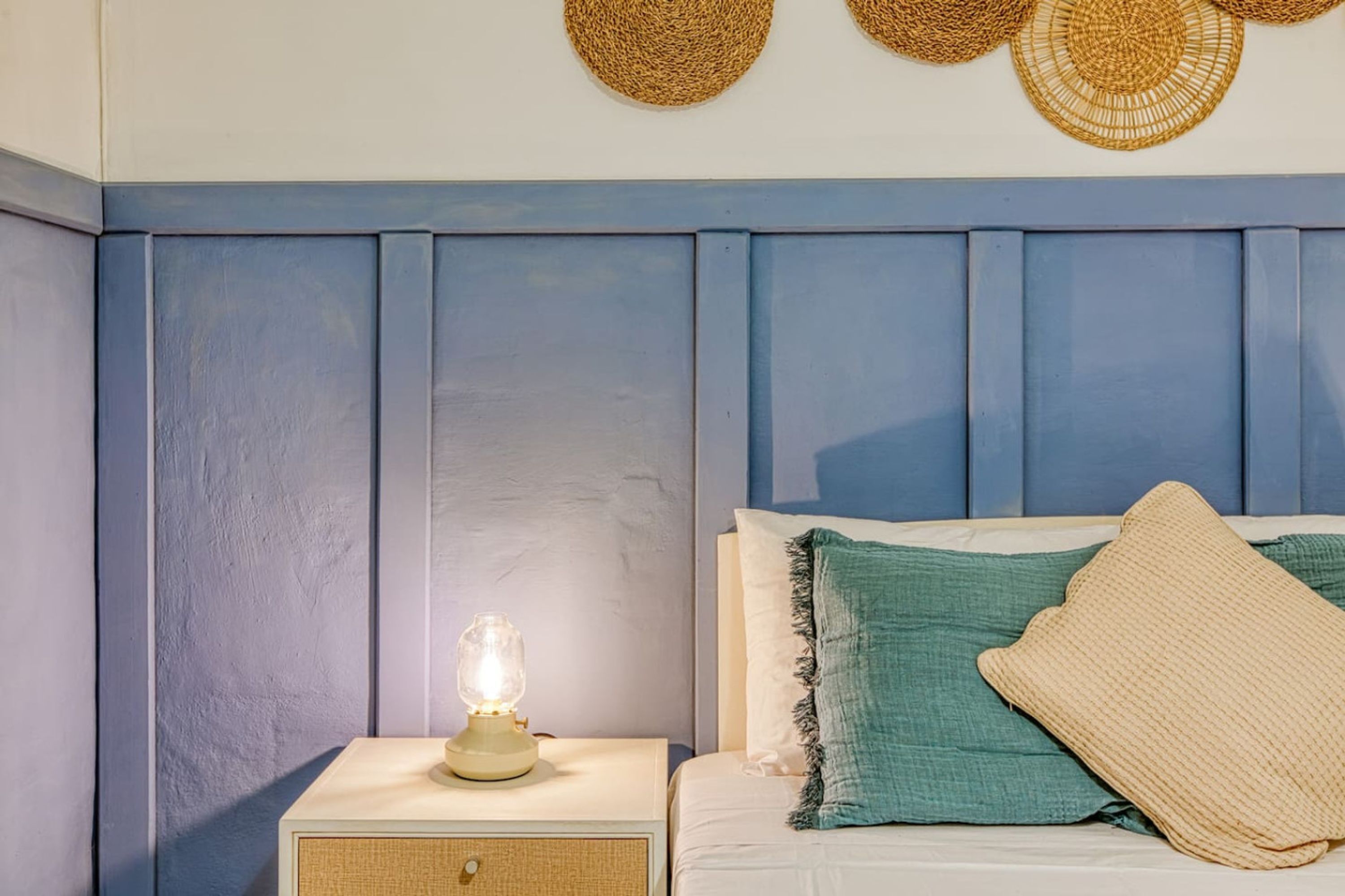 Ein Schlafzimmer mit blauem Wandpaneel als Kopfteil, einer Nachttischlampe und einem Bett mit grün-beiger Bettwäsche.