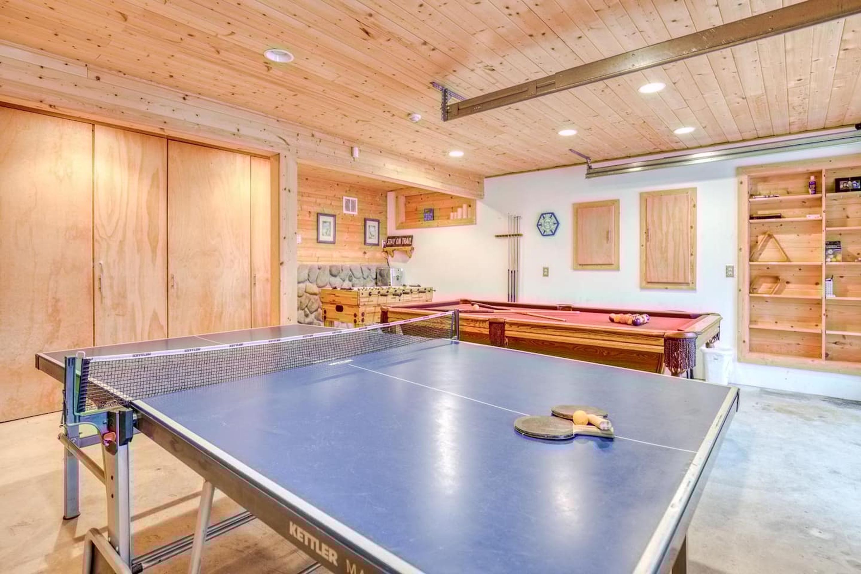 Ein Tischtennistisch in einem Raum mit Holzwänden und -decke, ein Billardtisch und Regale mit diversen Gegenständen.