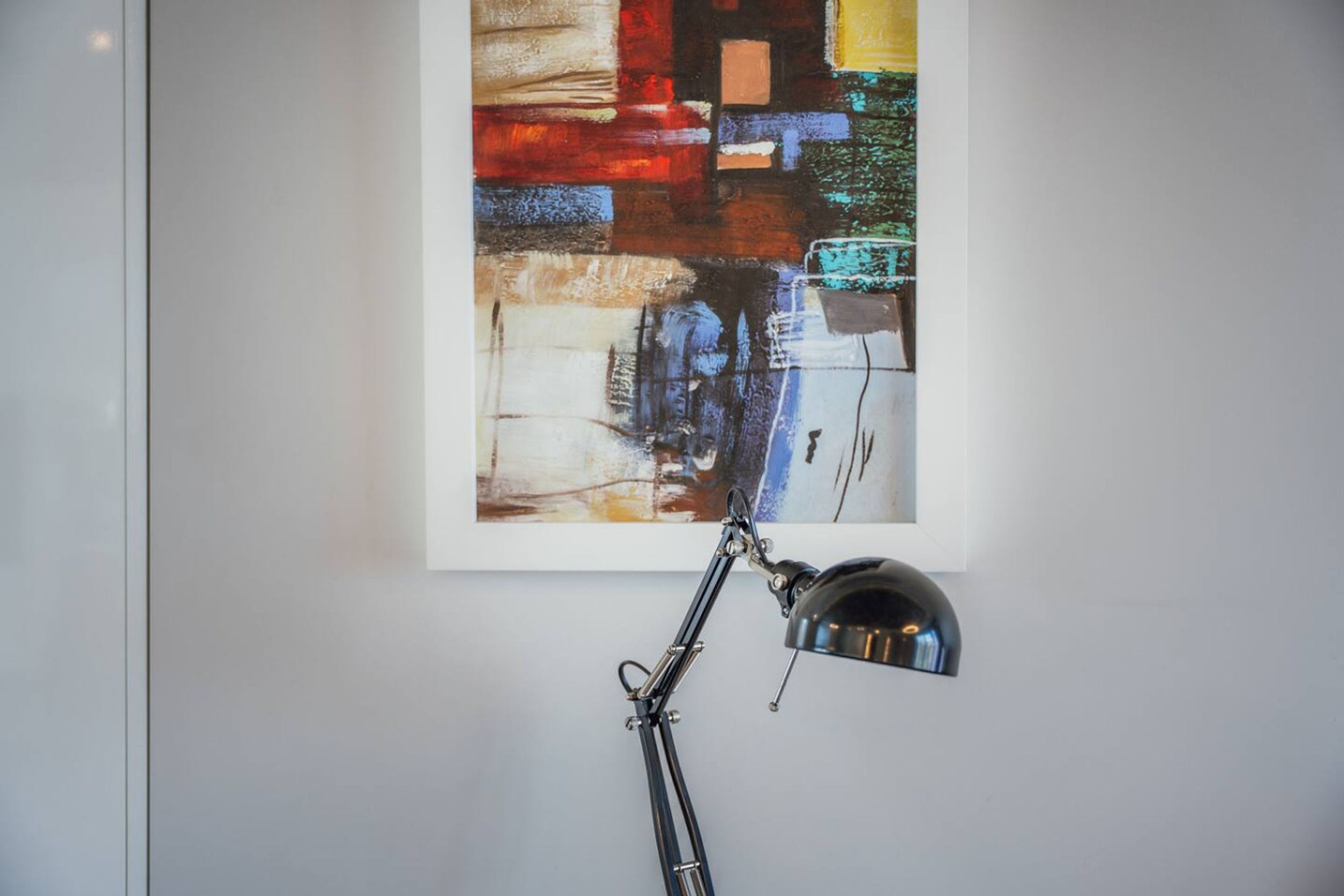 A framed abstract painting on a white wall with a black desk lamp in front.