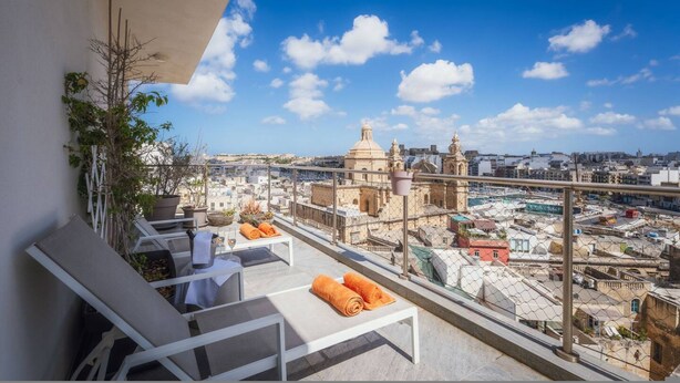 A rooftop terrace with white lounge chairs, orange towels, and a view of a historic cityscape with domed buildings. - Balanzone s Creative Penthouse (Msida)
