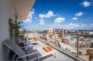 Une terrasse sur le toit, dotée de chaises longues blanches, de serviettes orange et offrant une vue sur un paysage urbain historique, avec ses bâtiments coiffés de dômes.