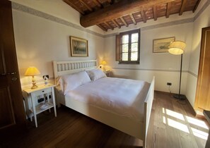 A bedroom with a wooden floor, a bed with white bedding, two bedside tables with lamps, a window, and a picture on the wall. - Agriturismo Borgo Sette Tigli Apt 5 (Montepulciano)