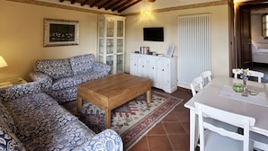 A living room with a patterned sofa, a wooden coffee table, a cabinet with glass doors, a television, and a dining area with a table and chairs. - Agriturismo Borgo Sette Tigli Apt 5 (Montepulciano)