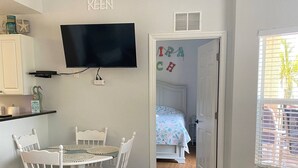 Een kamer met een eettafel en stoelen, een aan de muur bevestigde televisie, en een bed met kleurrijke letters die 'IRACHY KEEN' vormen.