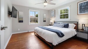 Une chambre à coucher avec un lit, un ventilateur de plafond, deux fenêtres et des œuvres d’art accrochées aux murs.