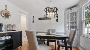 Un coin repas avec une table en bois, des chaises à l’assise rembourrée de couleur grise, un buffet noir contenant de l’ustensile de cuisine et une enseigne suspendue.