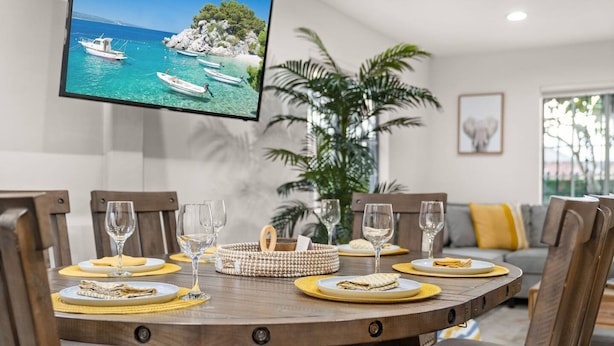 A dining area with a round wooden table set for four, surrounded by chairs. A flat-screen TV mounted on the wall displays a scenic coastal view.