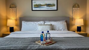 Ein Hotelzimmer mit einem großen Bett, zwei Nachttischlampen, einem gerahmten Landschaftsbild und einem Tablett mit Flaschen und Gläsern auf dem Bett.