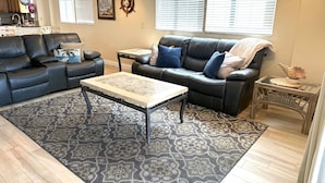 A living room with a dark leather sofa, a coffee table, and a patterned rug. There are two windows with blinds, a chandelier, and a small table with a decorative shell. - The Old Man the Sea-seaview 101 (Madeira Beach)