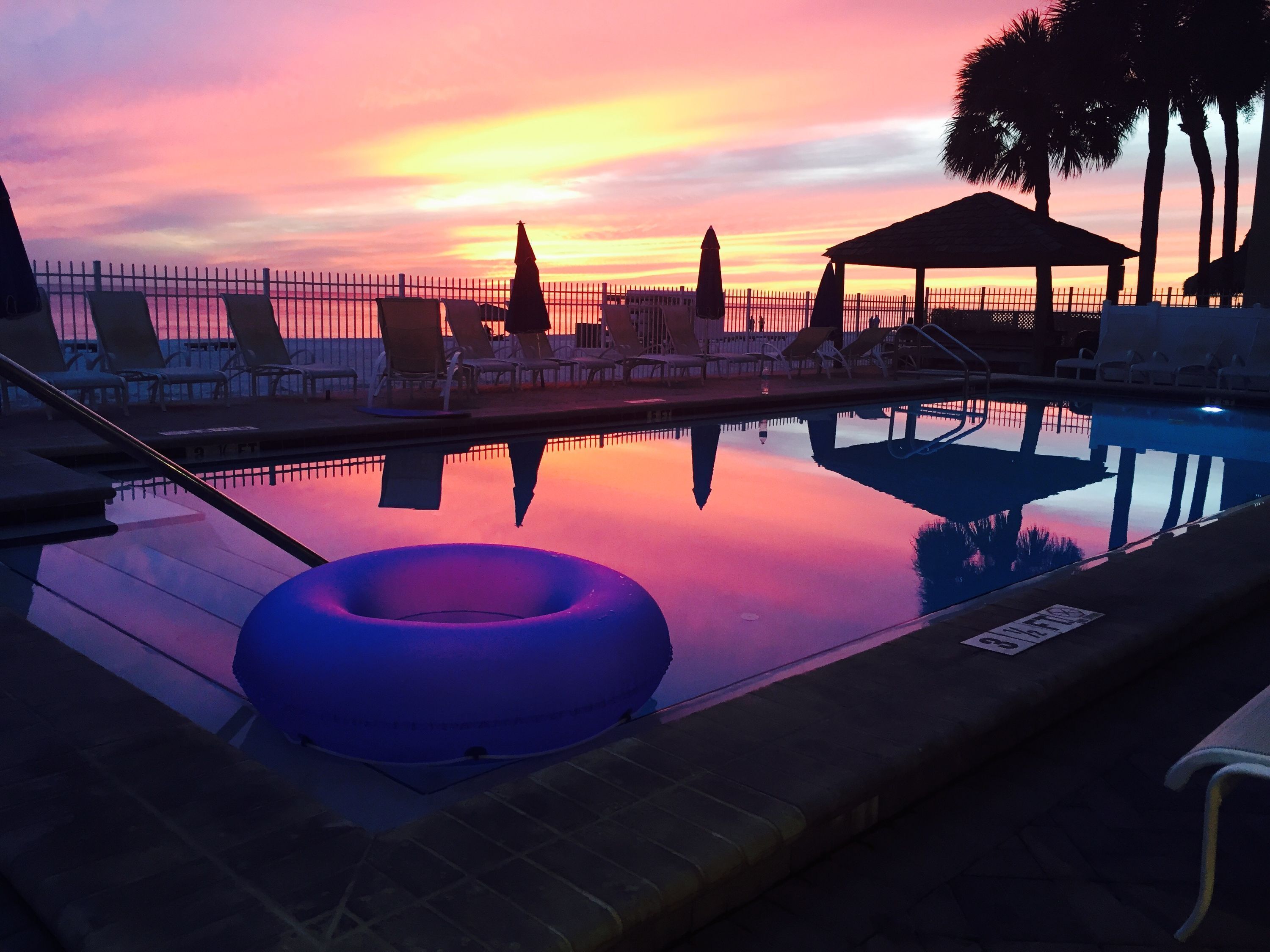 A swimming pool with lounge chairs and a gazebo, reflecting a sunset.