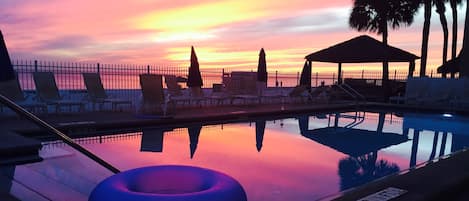 A swimming pool with lounge chairs and a gazebo, reflecting a sunset.