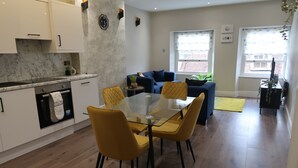 A modern kitchen with white cabinets and a marble countertop, a dining area with yellow chairs and a glass table, and a living area with a blue sofa and a green plant. - Casa Bella in Sherborne s Historic Quarter (Sherborne)