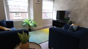 A living room with two dark blue sofas, a small round table with a plant and headphones, a green rug, and a TV on a black stand. - Casa Bella in Sherborne s Historic Quarter (Sherborne)