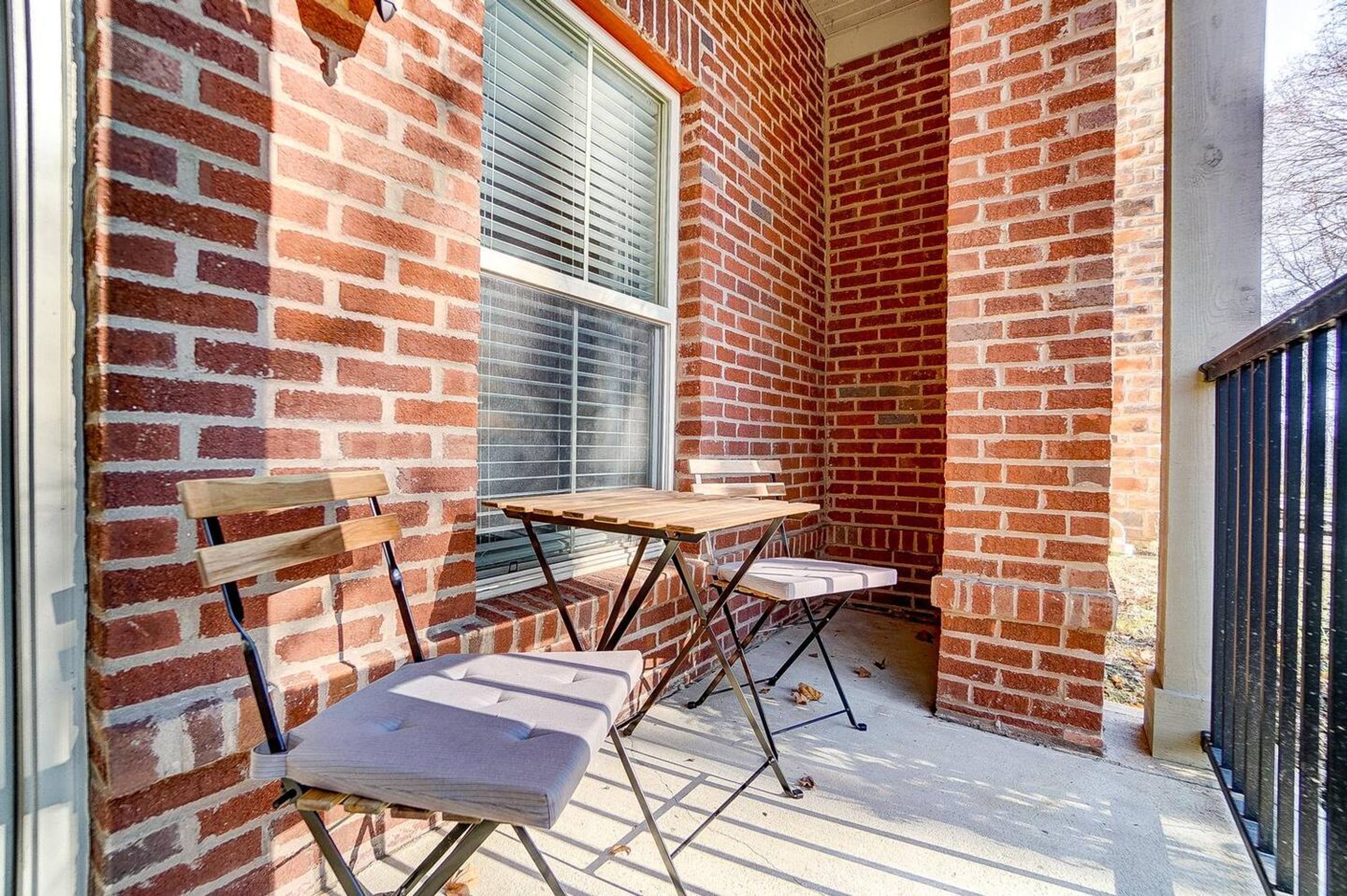 Une petite terrasse extérieure avec un mur en briques, une table en bois, deux chaises et une fenêtre avec des stores.