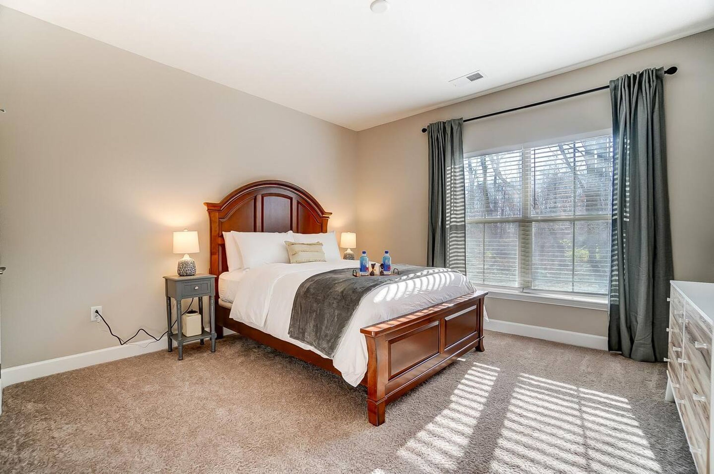 A bedroom with a wooden bed, two bedside tables with lamps, a window with curtains, and a view of trees outside.