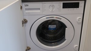 Una lavadora compacta en un pequeño espacio de cocina con encimera de mármol, un lavamanos y un hervidor.
